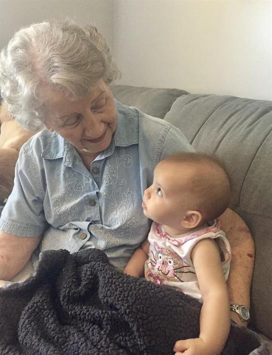 An elderly woman smiles as she interacts with a curious baby on a cozy sofa at home.