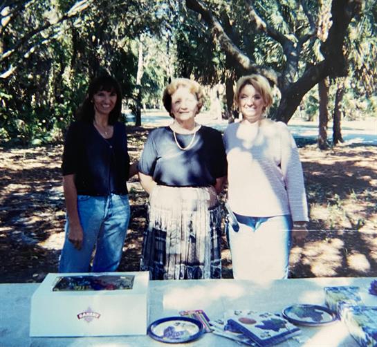 Three women stand together smiling at a park event, enjoying each other's company in nature.