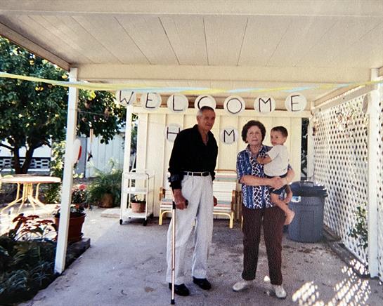 A grandchild is embraced by grandparents under a cheerful welcome banner at their home.