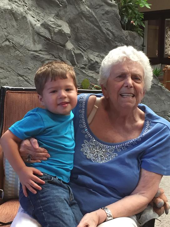 A joyful child sits on his grandmother's lap, both smiling in a warm and sunny outdoor space.