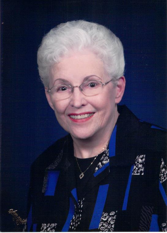 A senior woman with white hair and glasses smiles warmly in a formal setting.