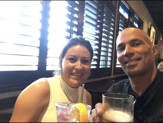 Two friends sit at a table holding drinks, smiling warmly while sunlight streams through blinds.