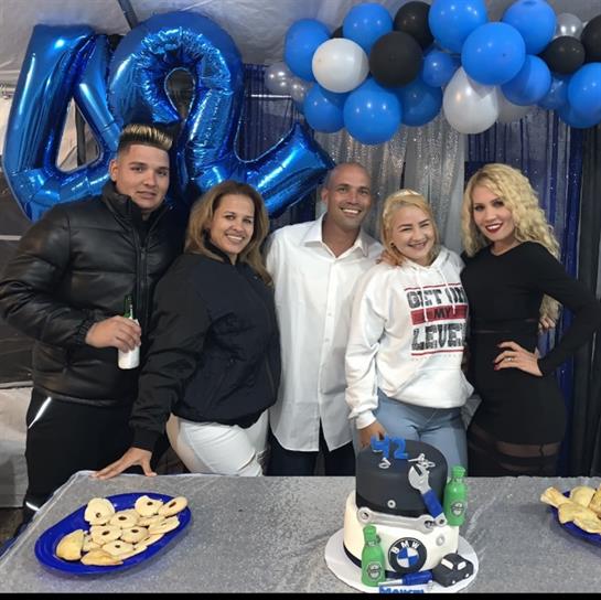 Friends celebrate indoors, smiling with a cake and balloons.