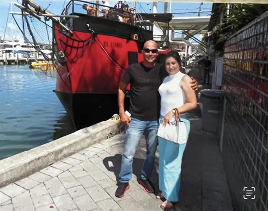 A couple stands smiling on a sidewalk by a vibrant red ship near the water on a sunny day.