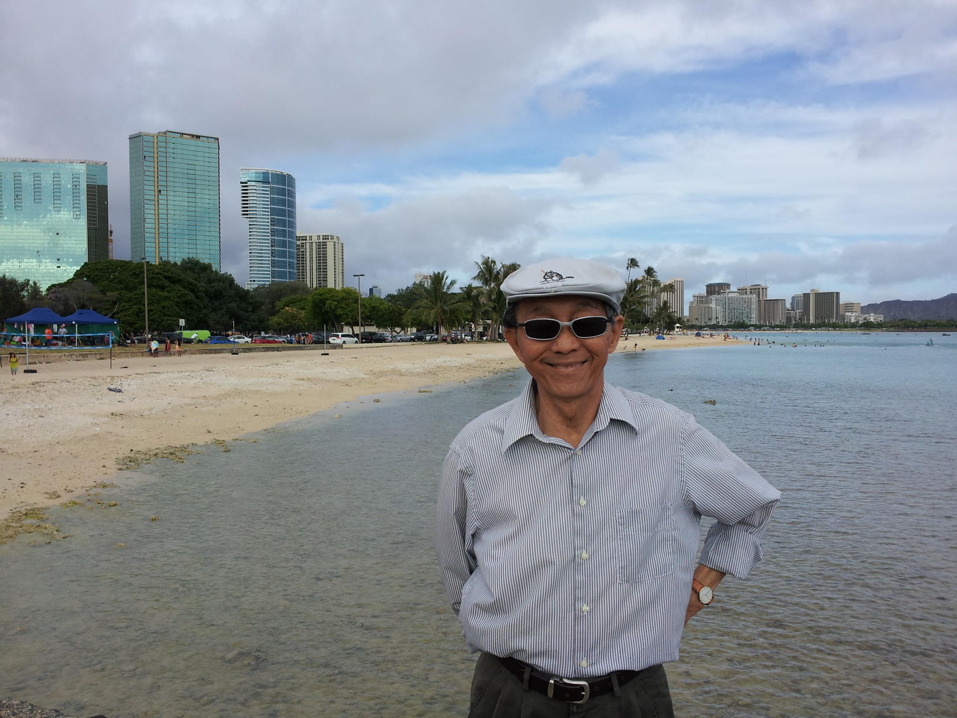 Elderly man in sunglasses stands by a beach with a city skyline and water behind him.