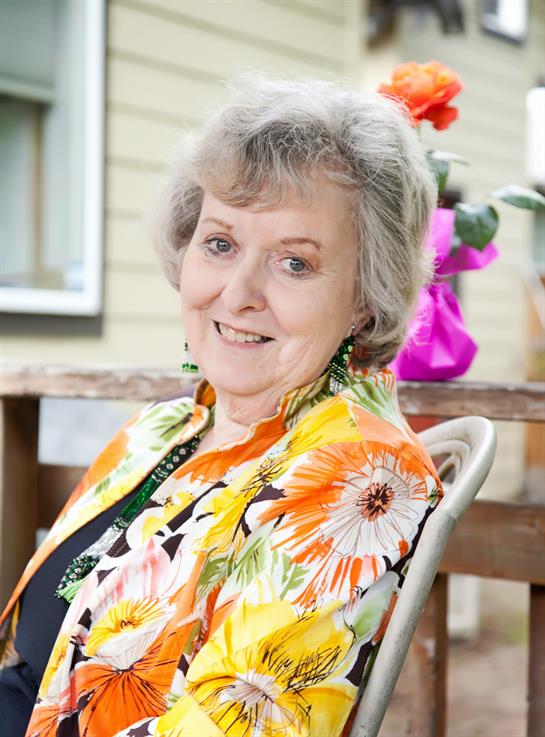 A woman with gray hair enjoys a warm day seated on a patio, wearing a vibrant floral jacket.