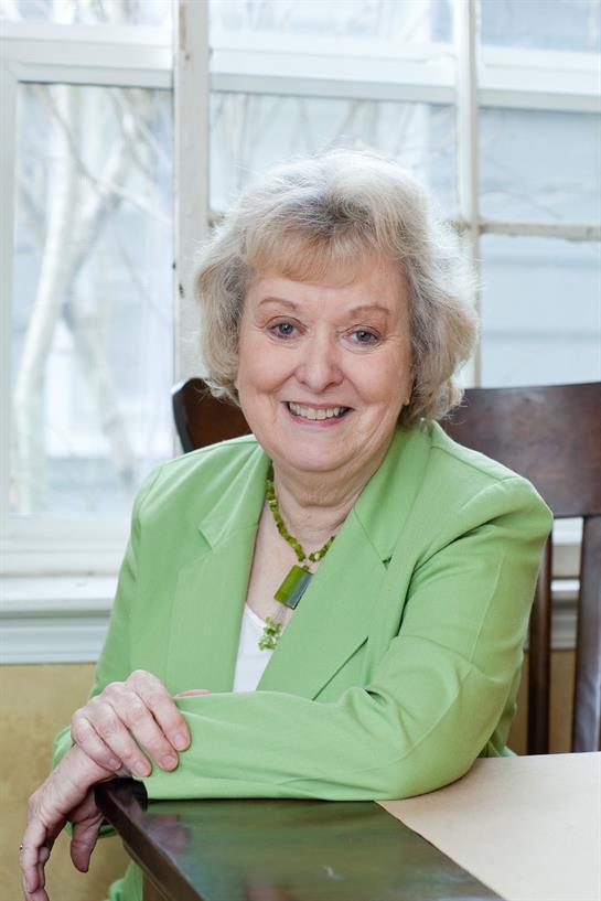 A cheerful older woman with curly hair smiles while sitting at a wooden chair near a window.