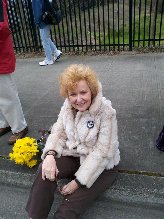 A smiling elderly woman sits on the sidewalk beside yellow flowers, enjoying a community gathering.