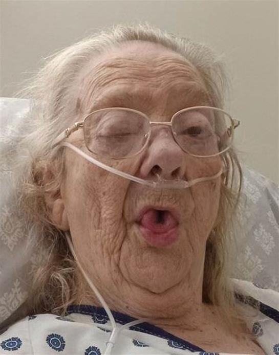 An elderly woman in a hospital bed playfully sticks out her tongue while wearing an oxygen mask.