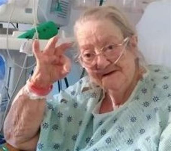 Woman in a hospital gown smiles and waves while resting in a healthcare facility room.