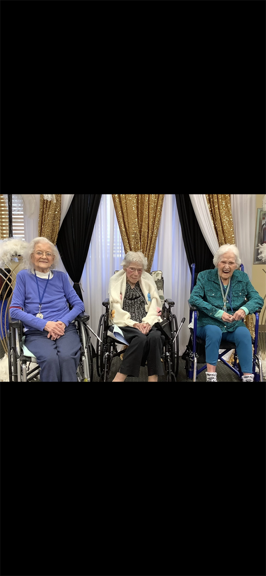Three elderly women sit side by side in wheelchairs, smiling and enjoying each other's company.