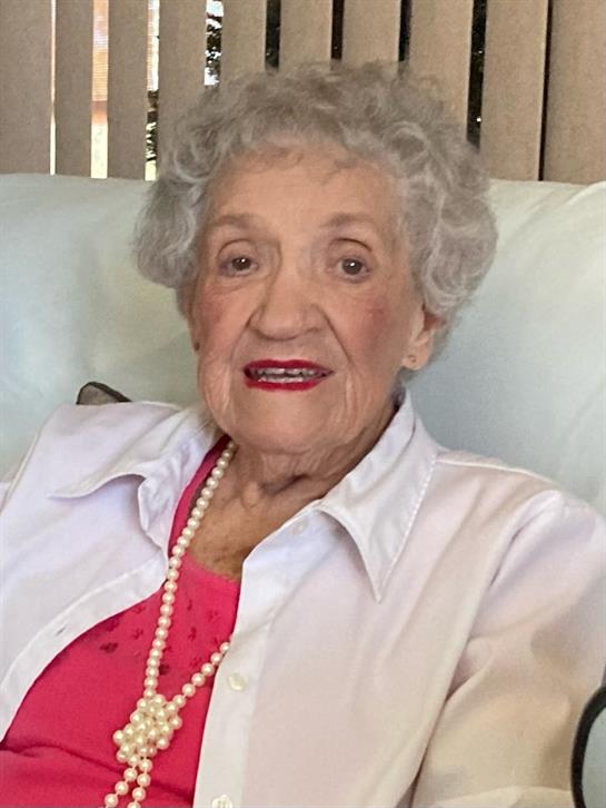An elderly woman with curly gray hair and red lipstick sits on a sofa, looking cheerful and relaxed.