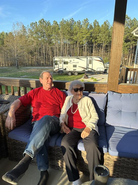 A cheerful couple relaxes on a cozy porch while surrounded by tall trees and a camper.
