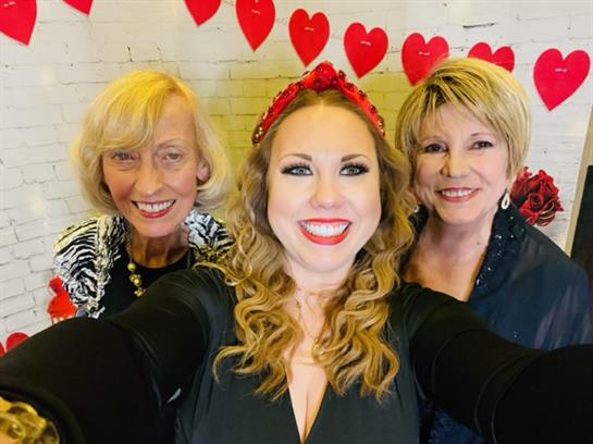 Three friends pose happily for a selfie at a decorated venue filled with heart shapes.