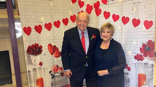An elderly couple dressed elegantly stands in front of heart-themed decorations for a celebration.