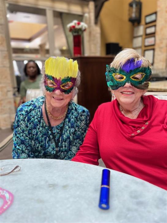 Two cheerful women wear vibrant masks and smile at a festive gathering in a bright location.