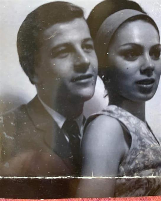 A young couple in formal attire stands close, smiling against a plain background.