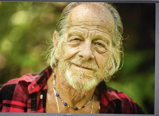 An elderly man with a gentle expression poses outdoors, surrounded by greenery and natural light.