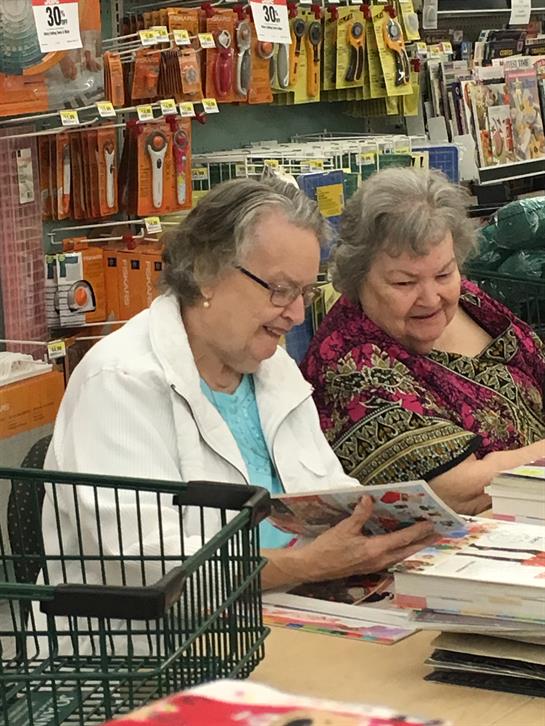 Two older women smile and chat while browsing craft supplies together.