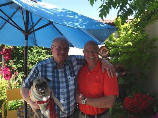 Two men stand in a vibrant garden, smiling while holding a pug under a blue umbrella.
