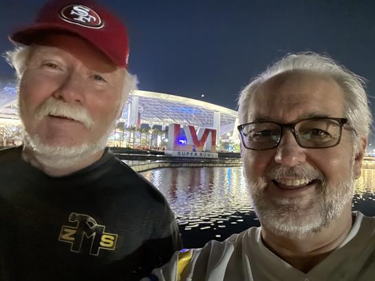 Two smiling friends take a selfie by the water at night, with bright stadium lights behind them.