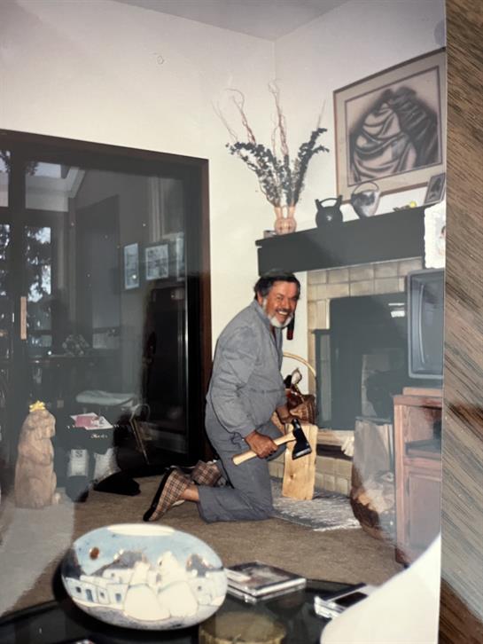 A man kneels by the fireplace, chopping firewood with enthusiasm in a cozy living room setting.