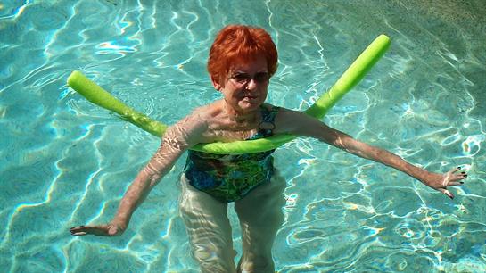 A senior woman participates in water aerobics using a pool noodle in a vibrant blue pool.