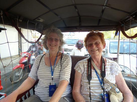 Two friendly women sit together in a rickshaw, smiling as they ride through a lively street.