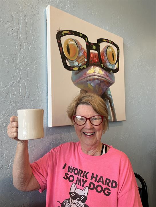 A cheerful elderly woman raises a coffee mug, smiling in front of unique frog artwork at a cafe.