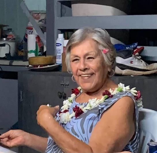 A joyful elderly woman in a floral necklace sits indoors, happily engaging with others.