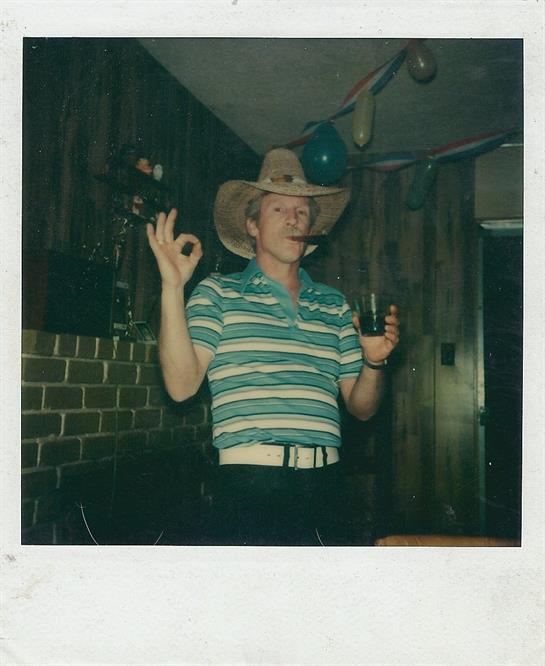 A man with a cowboy hat and striped shirt stands cheerfully holding a drink in a wooden interiors.