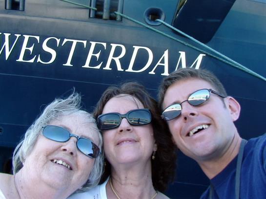 Three people pose happily together by a large cruise ship on a clear day, smiling widely.