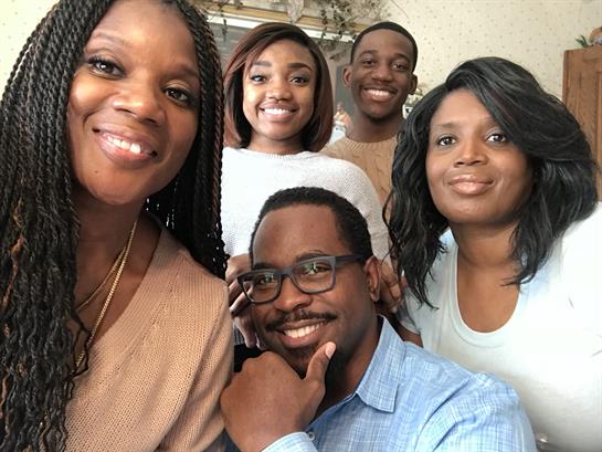 Five family members smile and pose together in a bright, cozy living room, enjoying their time.