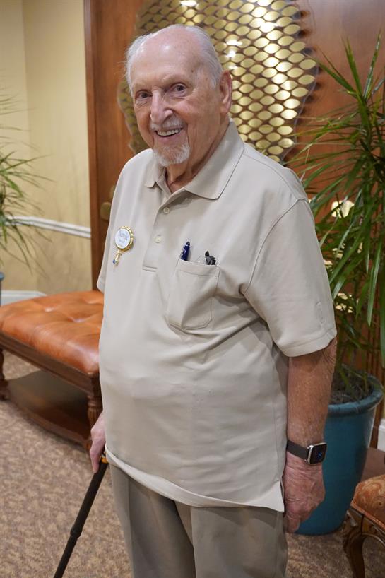 Elderly man with a friendly expression stands indoors surrounded by decorative plants and furniture.