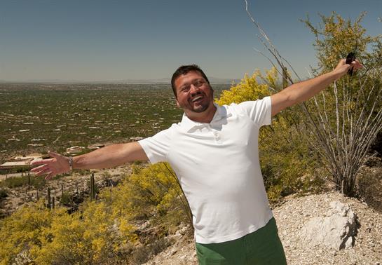 A man smiles widely while standing on a mountain, arms open to embrace the scenic view below.