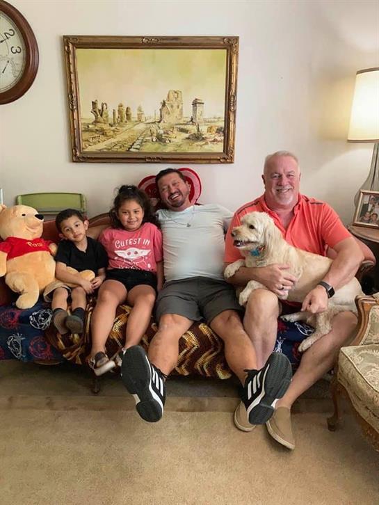 Adults and children relax on a couch surrounded by toys and a dog in a warm living room setting.