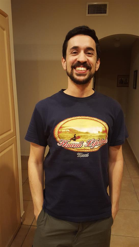 A man stands in a hallway, smiling while wearing a t-shirt featuring a scenic sunset design.