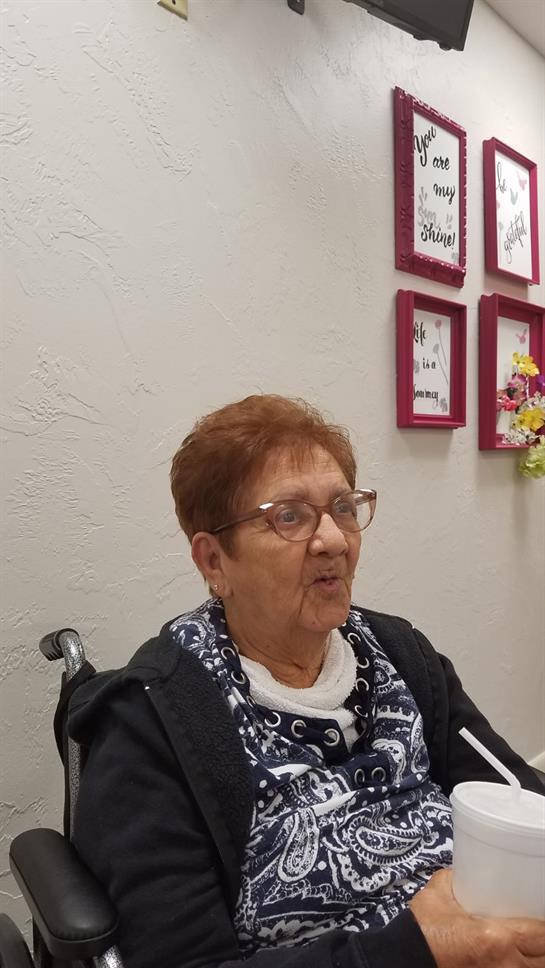 An elderly woman engages in lively conversation while seated in a nursing home filled with decor.