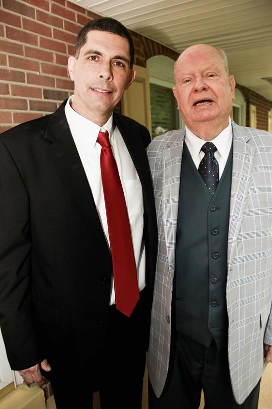 Two well-dressed men smile and stand side by side during a social gathering inside a building.