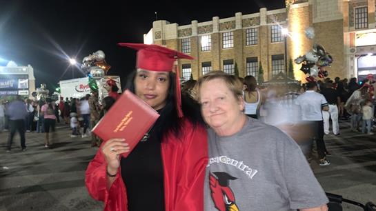 A graduate in a red cap and gown proudly holds her diploma while smiling with a family member.