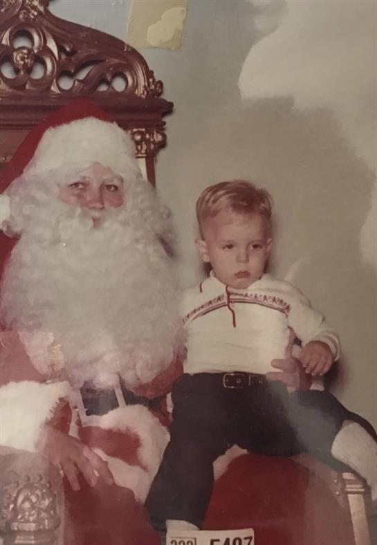 A young boy wearing a white shirt sits beside Santa Claus, both posing for a holiday moment.