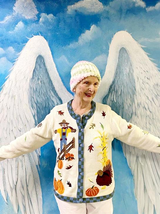 An elderly woman smiles brightly while standing with open arms against a vibrant angel wings mural.