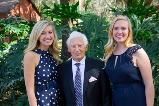 Two young women and an older man smile while standing among lush greenery in a garden.