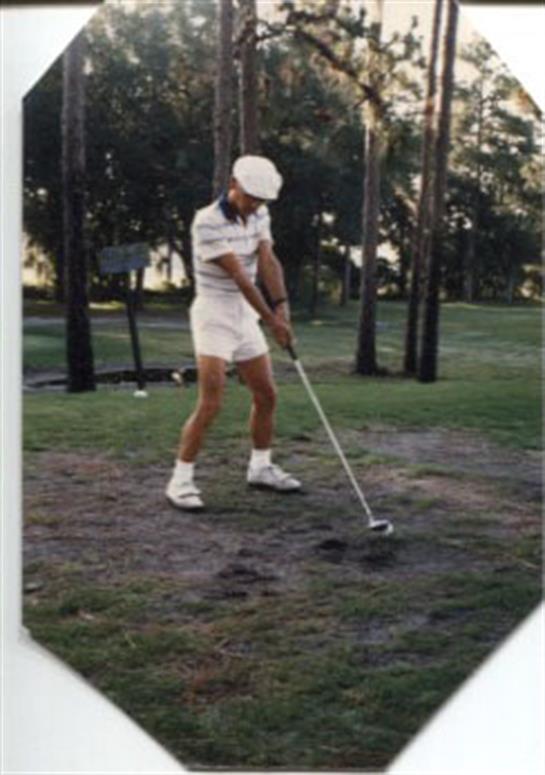 A man in a white outfit takes a golf swing in a lush green setting with tall trees nearby.