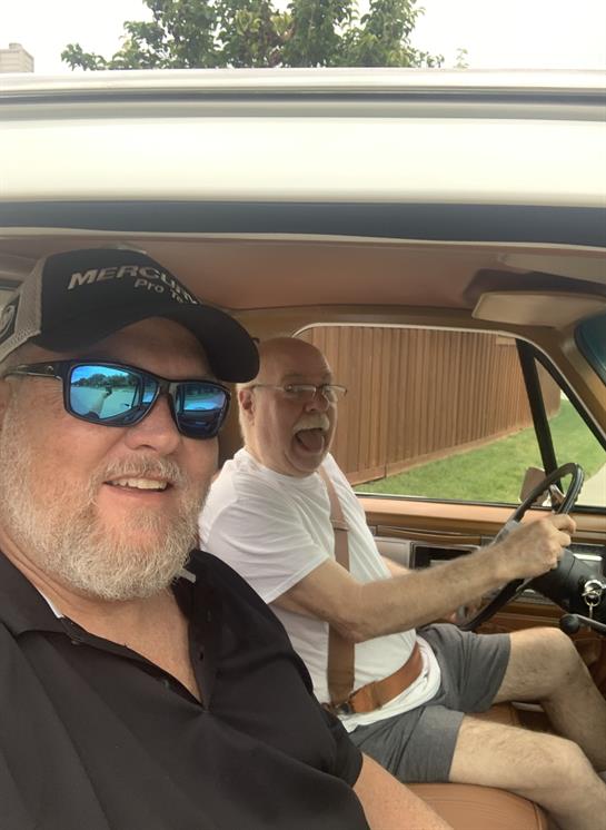 Two men smile while sitting inside a vintage car, one driving and the other enjoying the moment.