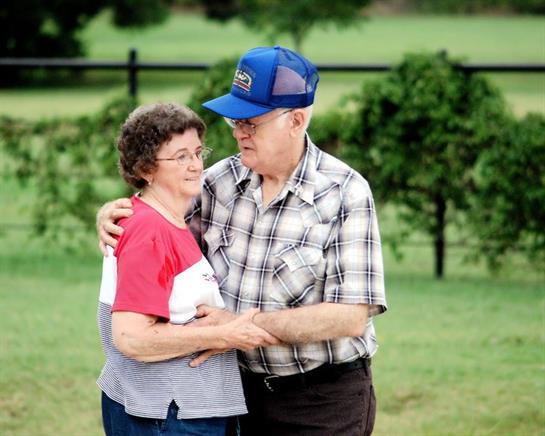 An elderly couple holds each other close while dancing in a beautiful garden, filled with greenery.