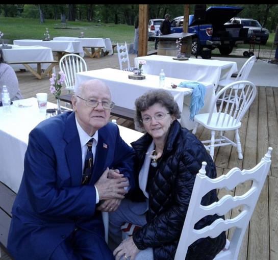 Couple sitting together at a table on a wooden deck surrounded by nature, smiling warmly.