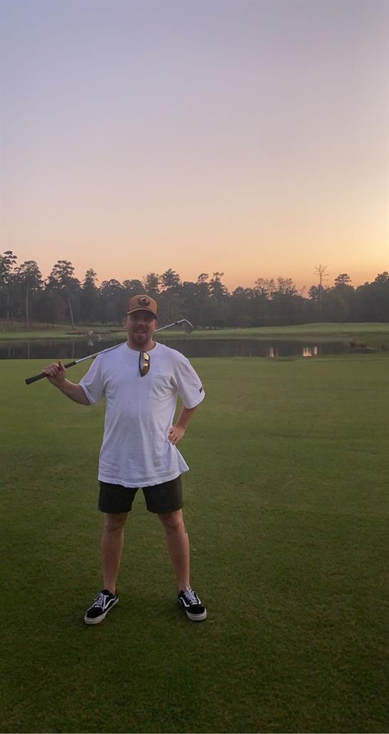 A man stands confidently on a golf course at sunset, club in hand, with a calm lake behind.