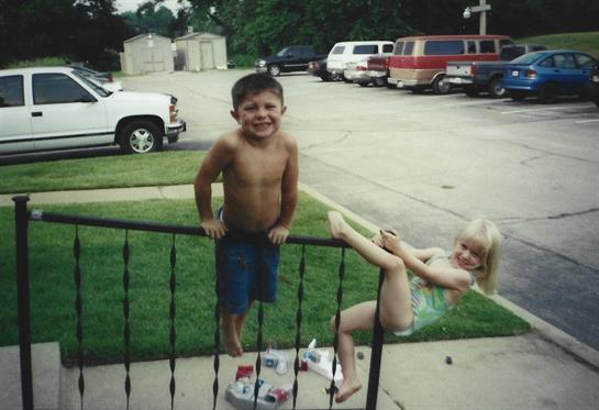Two kids play outside, one climbs a railing while the other sits nearby, enjoying summer fun.