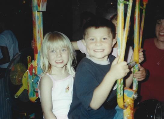 Two children joyfully ride a carousel at an amusement park, lit up in the evening.